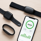 A flat lay of various fitness gadgets on a light wooden surface, including a black smartwatch, a sleek fitness tracker, a heart rate monitor, and a smartphone displaying fitness statistics. The items are arranged neatly